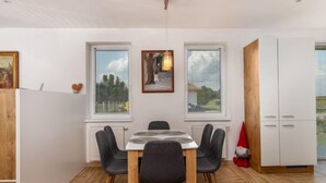 Un espace repas moderne avec une table et des chaises en bois, de grandes fenêtres équipées de stores et une vue sur l’extérieur.