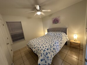 Une chambre à coucher comprenant un lit, un ventilateur de plafond, une fenêtre avec des stores, une table de chevet avec une lampe et un élément de décoration murale.