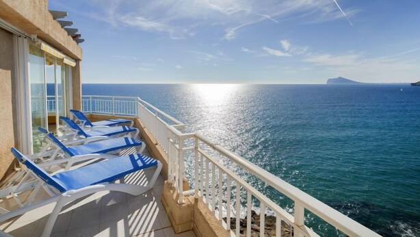 Ein Balkon mit blauen Liegestühlen mit Blick auf ein klares, blaues Meer unter strahlendem Sonnenschein.