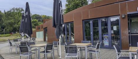 Un espacio al aire libre con mesas y sillas, una terraza de madera y un edificio moderno con puertas de vidrio.