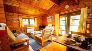 Una cabaña acogedora con paredes y suelos de madera, un sofá, una mesa de comedor con sillas, un televisor y un ventanal con cortinas.
