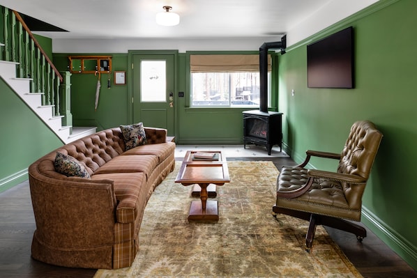 A living room with a brown leather sofa, a wooden coffee table, and a leather armchair. A staircase with green walls and a wooden railing is visible.
