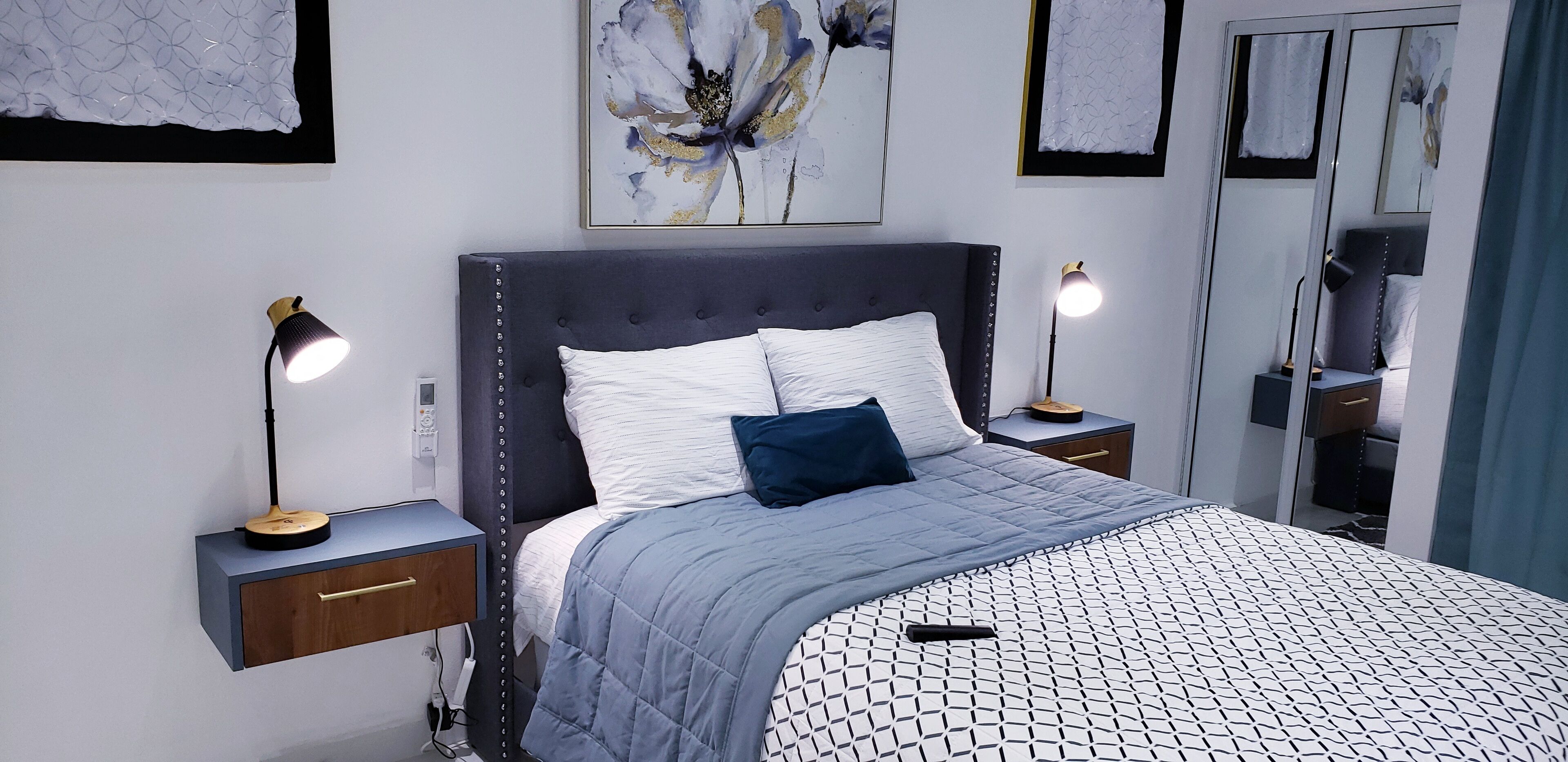 A modern bedroom with a tufted headboard, a grey quilted bedspread, two bedside lamps, a nightstand with a drawer, and a large mirror on the wall.