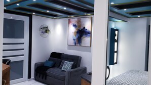 Une pièce moderne avec un canapé gris, une petite table, une étagère murale décorée de divers objets, et un grand tableau abstrait.