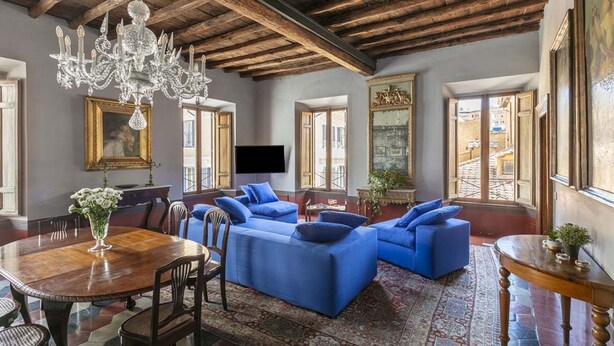 Un salón espacioso con techo de madera, una araña, sofás azules, una mesa de comedor y un televisor.