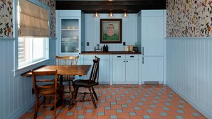 A kitchen with a dining table, chairs, a window with blinds, a framed picture on the wall, and patterned wallpaper.