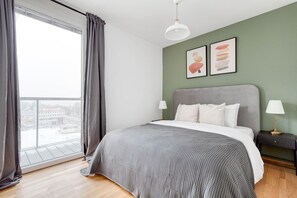 Quarto moderno com cama grande, cortinas cinzas, vista da cidade e duas obras de arte emolduradas na parede.