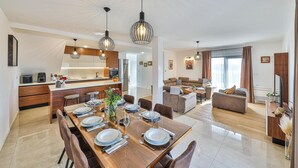 Una cucina moderna con isola centrale, zona pranzo con tavolo in legno apparecchiato per sei e salotto con divani e televisore.