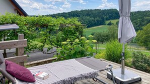 Eine Holzterrasse mit gepolstertem Stuhl, einem kleinen Tisch mit einem Buch und einem Sonnenschirm.