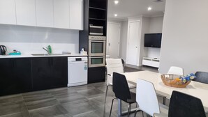 Una cocina moderna con armarios blancos y negros, horno empotrado y lavavajillas. Hay un comedor con mesa y sillas, y un televisor montado en la pared.