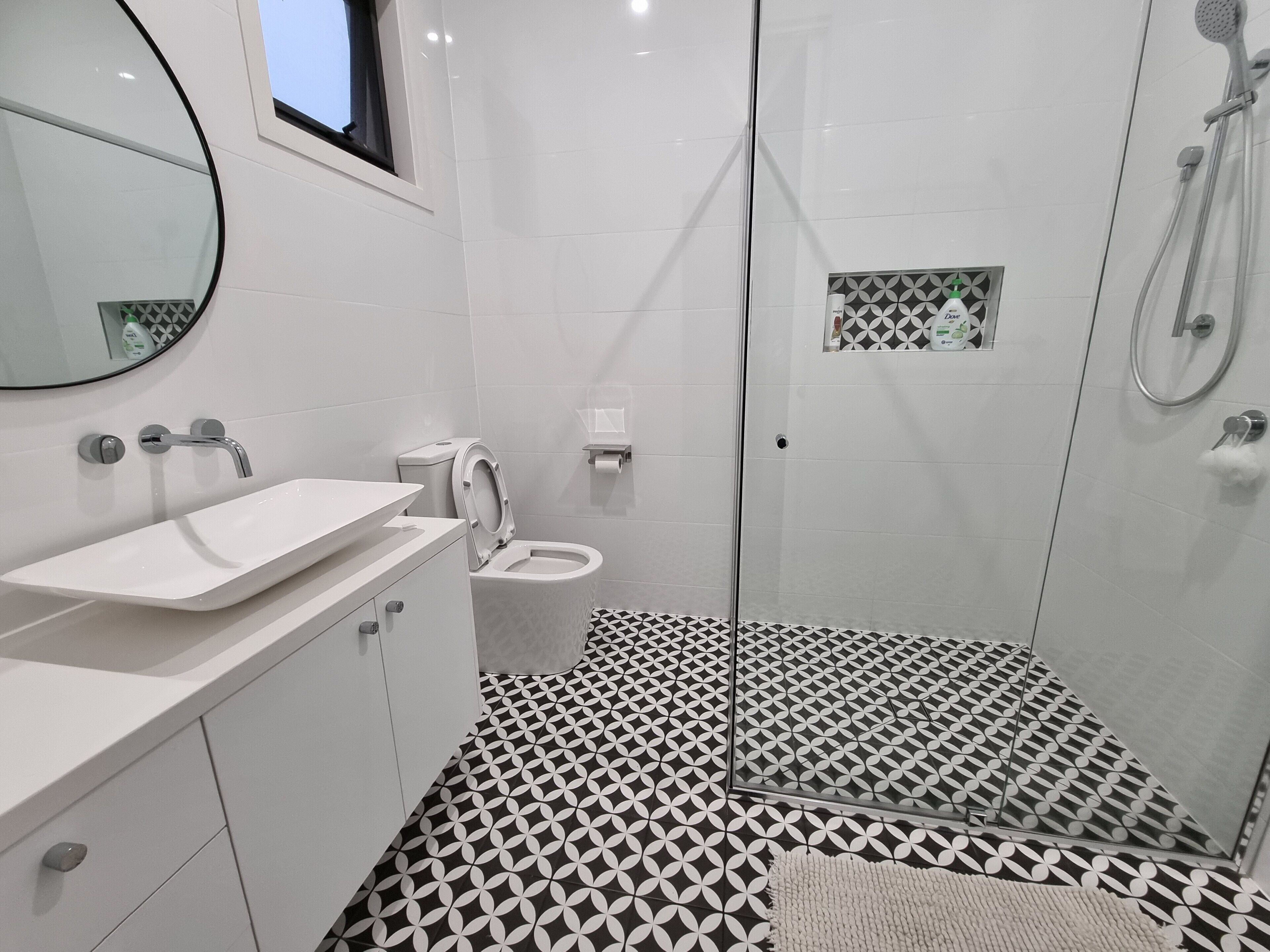 A modern bathroom with a white sink, toilet, and shower. The floor has a black and white geometric tile pattern.