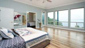 Un dormitorio amplio con una cama grande, ventilador de techo y vista al mar a través de ventanas amplias.