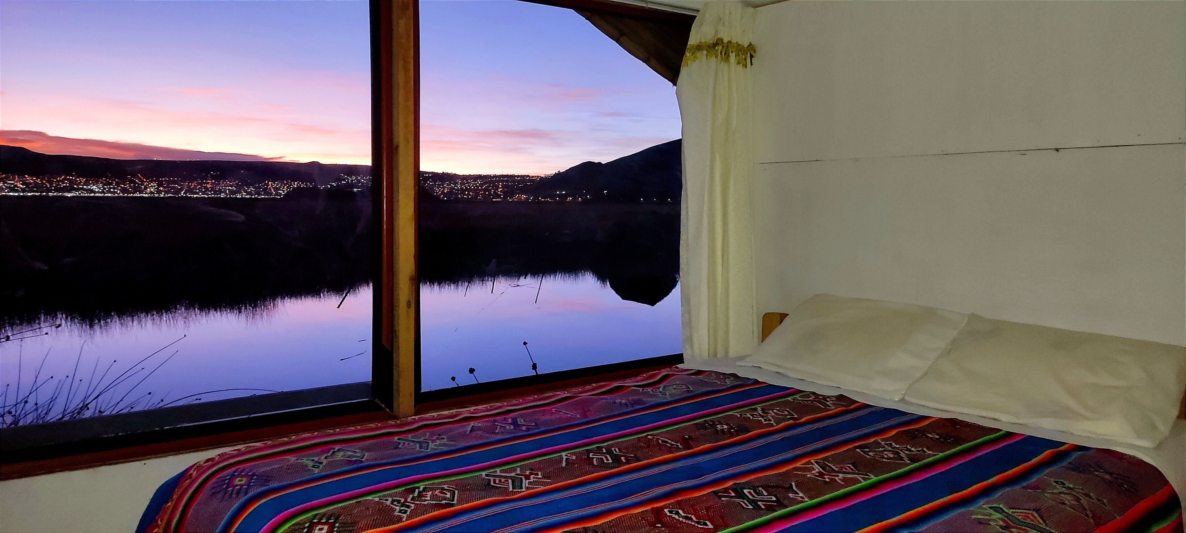 A room with a large window overlooking a lake and city lights at dusk.