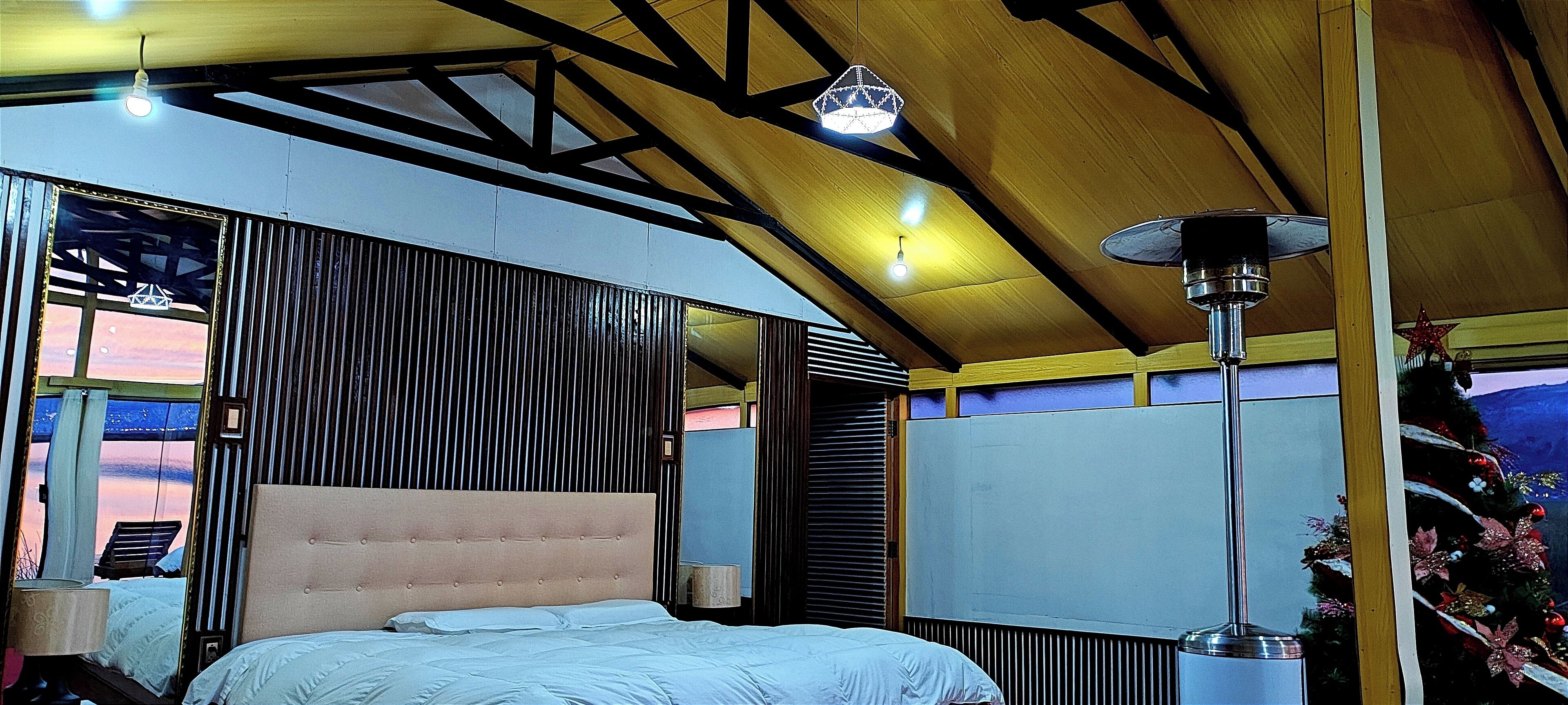 Une chambre à coucher avec un lit, une table de chevet, une lampe, une fenêtre avec des rideaux et une vue sur le coucher de soleil.