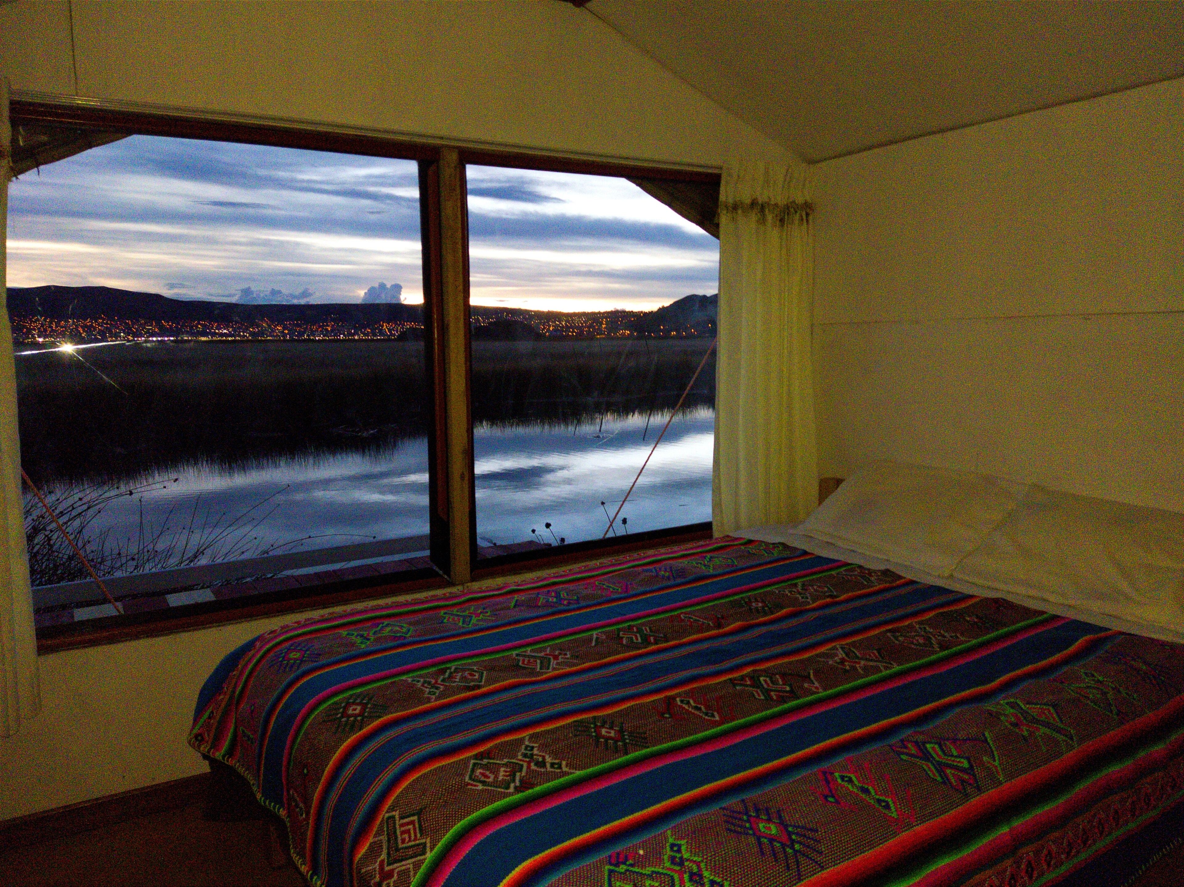 A bedroom with a large window offering a view of a lake and city lights at dusk.