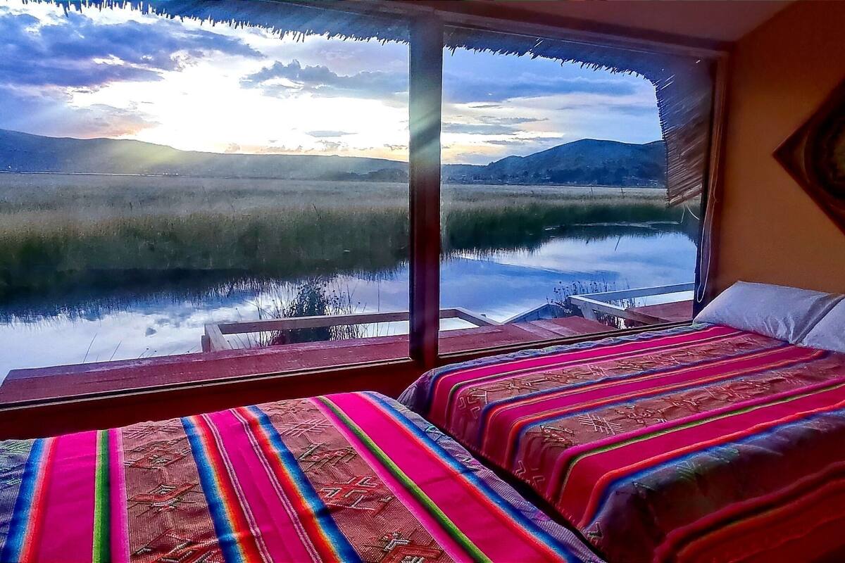 A room with a large window overlooking a lake and mountains, two beds with colorful bedding, and a wooden bench.
