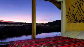 A room with a large window overlooking a cityscape at dusk, a bed with a patterned blanket, and a wall with a lotus design.