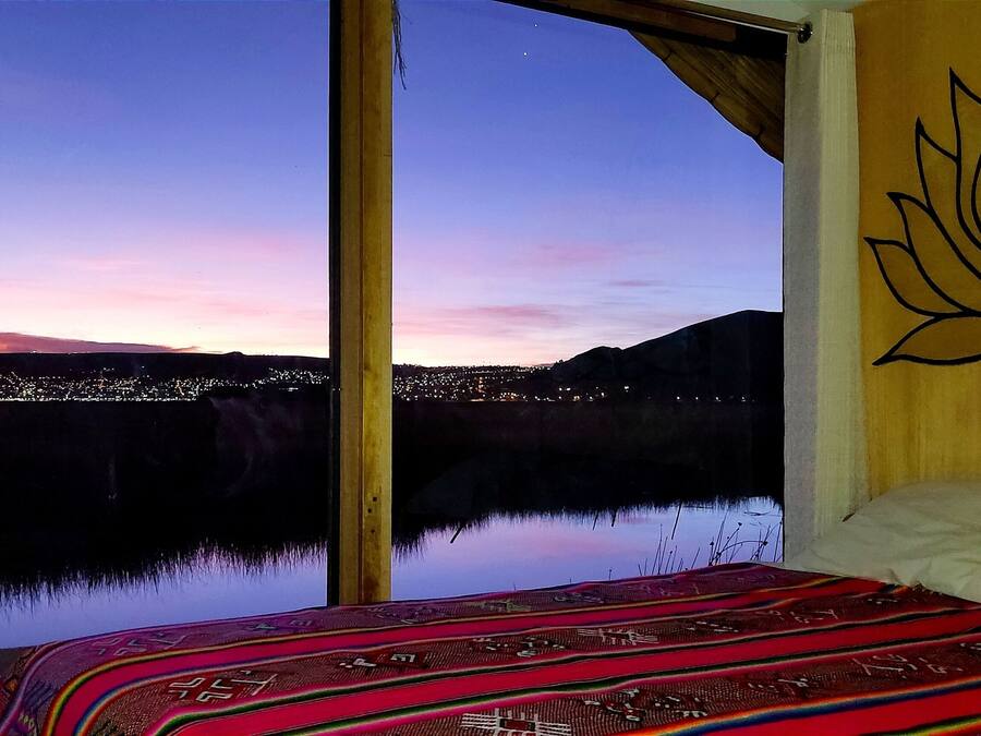 A room with a large window overlooking a cityscape at dusk, a bed with a patterned blanket, and a wall with a lotus design.