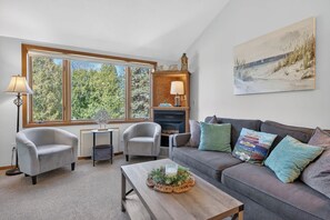 Un salon avec une cheminée, deux fauteuils, un canapé agrémenté de coussins colorés, une table basse avec un élément décoratif central et un tableau accroché au mur.