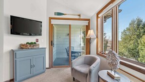 A room with a flat-screen TV, a grey sofa, a small table with a lamp, and a large window with a view of trees. - Ephraim Condo | Walk to Water, Wilson's and Shops (Ephraim)