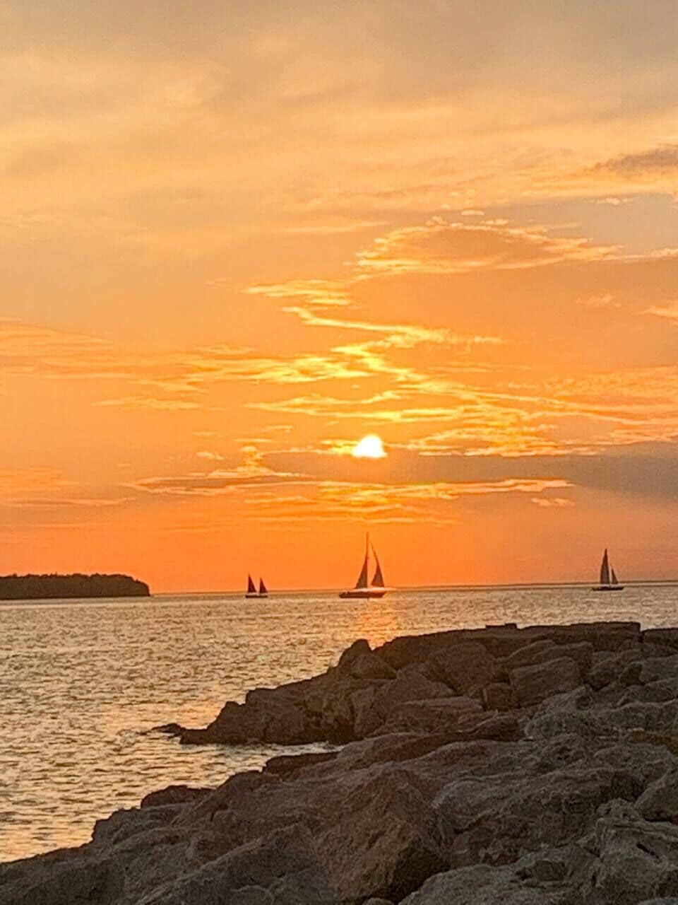A sunset over a rocky shoreline with sailboats in the distance.