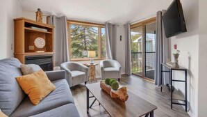 A living room with a sofa, two armchairs, a coffee table, a wooden bookshelf, a TV mounted on the wall, and a sliding glass door leading to a balcony. - Ephraim’s Pearl (Unit 203) | Balcony Views, Walk to Waterfront and Downtown Charm (Ephraim)