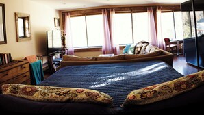 A bedroom with a large bed, a desk, and a chair. There is a window with curtains, a dresser, and a television.