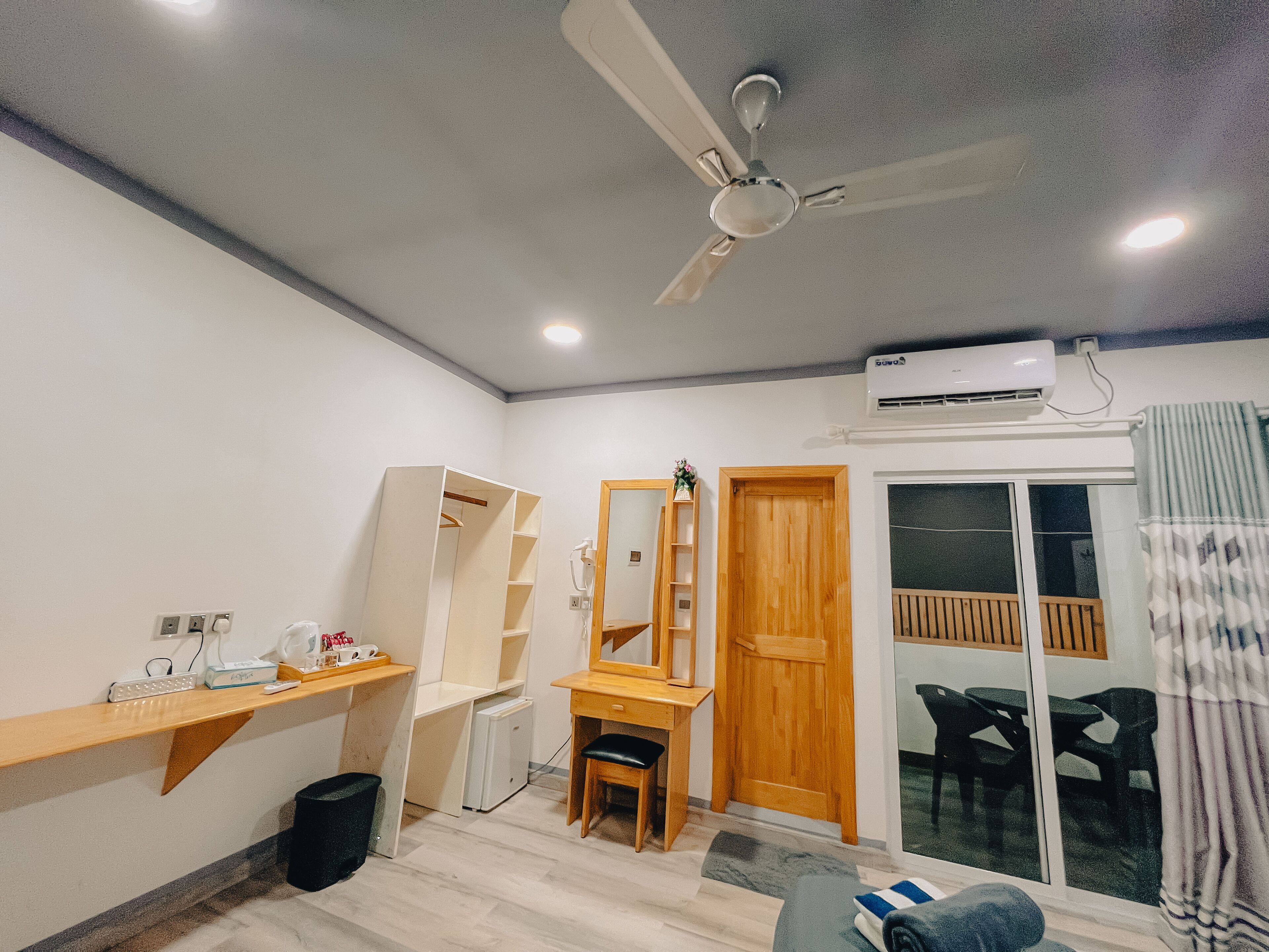 Una habitación bien iluminada con ventilador de techo, un escritorio con silla, un refrigerador pequeño, una puerta de madera y una ventana con vista a muebles de exterior.