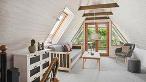 Un dormitorio acogedor en el ático, con techo inclinado, un sofá, una silla, una mesita y una ventana con vistas a la vegetación.