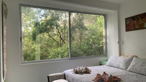 A bedroom with a large window, a bed with a patterned bedspread, a bench, and a painting on the wall. - Remarkable Studio in jungle of Chemuyil, Tulum (Chemuyil)