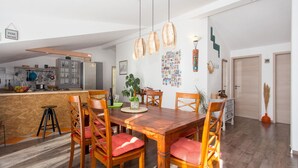Uma sala de jantar com mesa e cadeiras de madeira, uma cozinha ao fundo e uma parede com uma colagem de fotos.