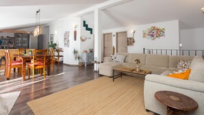 Uma sala de estar espaçosa com sofá, mesa de centro e área de jantar com cadeiras. Dá para ver uma cozinha ao fundo, com armários e geladeira.