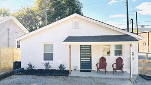 Una casita blanca con un porche, dos sillas de madera y un pequeño jardín.