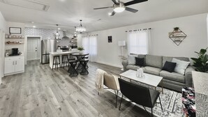 Una cocina moderna con una isla central, una zona de comedor con una mesa y sillas blancas, un sofá gris y un ventilador de techo.