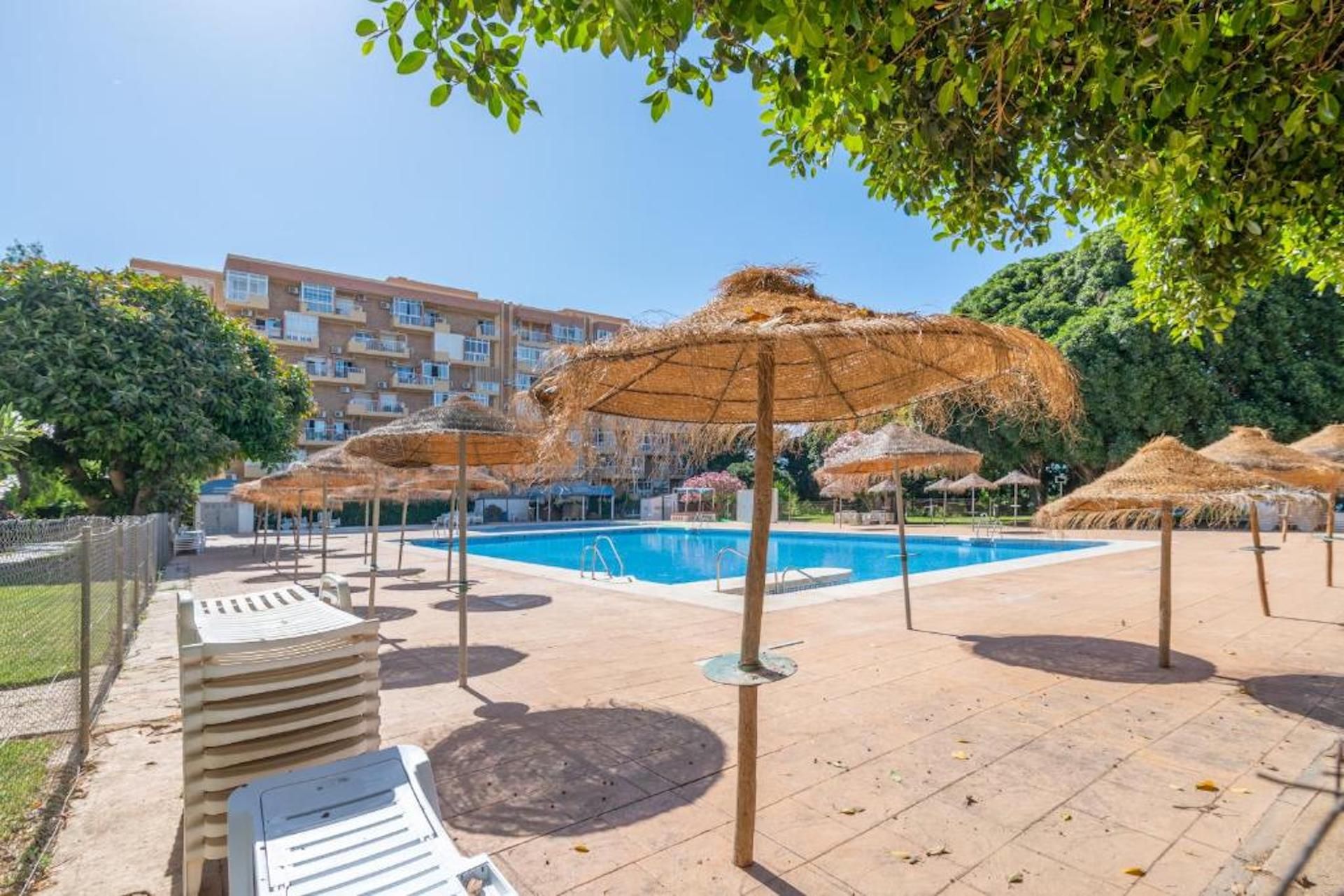 Un espace piscine agrémenté de parasols et de chaises longues, entouré d’un bâtiment doté de nombreux balcons.