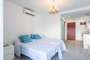 A bedroom with a bed, bedside tables, an air conditioning unit, a chandelier, and a view into the adjacent kitchen area.