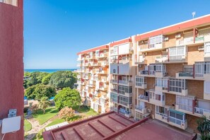 A multi-story residential building with balconies, a clear view of the sea, and a green landscape in the background.