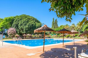 A swimming pool with a clear blue water surface, surrounded by lounge chairs and thatched umbrellas, set against a backdrop of lush greenery and a clear sky.