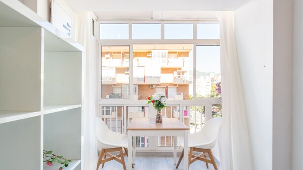 A balcony with a wooden table and two white chairs, a vase with flowers, and a view of neighboring buildings. - RENTAS Benalmádena Beach Studio (Benalmadena)