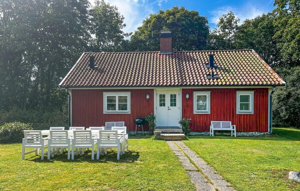 Una casa roja con techo de tejas, una puerta principal y dos ventanas. Hay un banco blanco y una mesa con sillas frente a la casa.