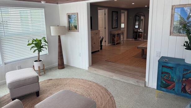 A spacious living room with a wooden ceiling, a large window with blinds, a floor lamp, a round rug, and a blue cabinet with decorative panels. - Beach house - 3 min walk to beach! (Englewood)