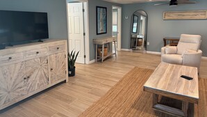 A modern living room with a TV on a wooden cabinet, a beige armchair, a coffee table, and a mirror on the wall. - Beach house - 3 min walk to beach! (Englewood)