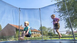 Dos niños saltando en un trampolín en un patio trasero con una casa y árboles al fondo.
