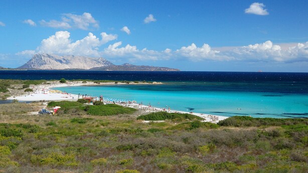 Een kustlandschap met een strand, helder turquoise water en een berg op de achtergrond.
