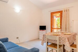 Una habitación con una mesa de comedor preparada para dos personas, un televisor sobre un mueble bajo, un sofá azul y una ventana con cortinas.