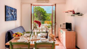 A dining area with a table set for two, a bowl of fruit, a bottle of wine, and a view of greenery outside.
