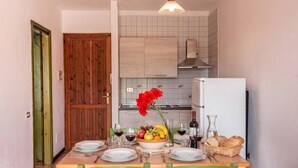 A dining area with a wooden table set for two, a bowl of fruit, and a bottle of wine.