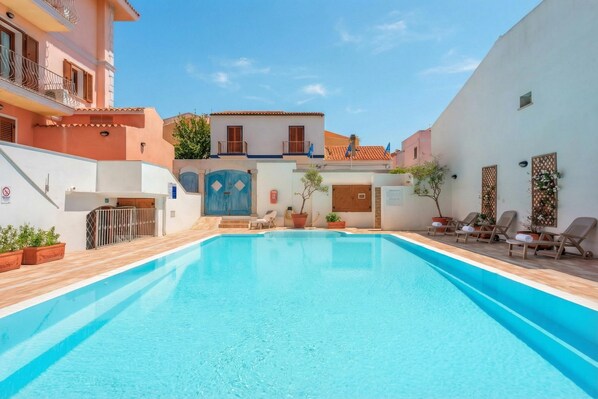 A swimming pool with a wooden deck, a blue gate, and a white building with balconies.