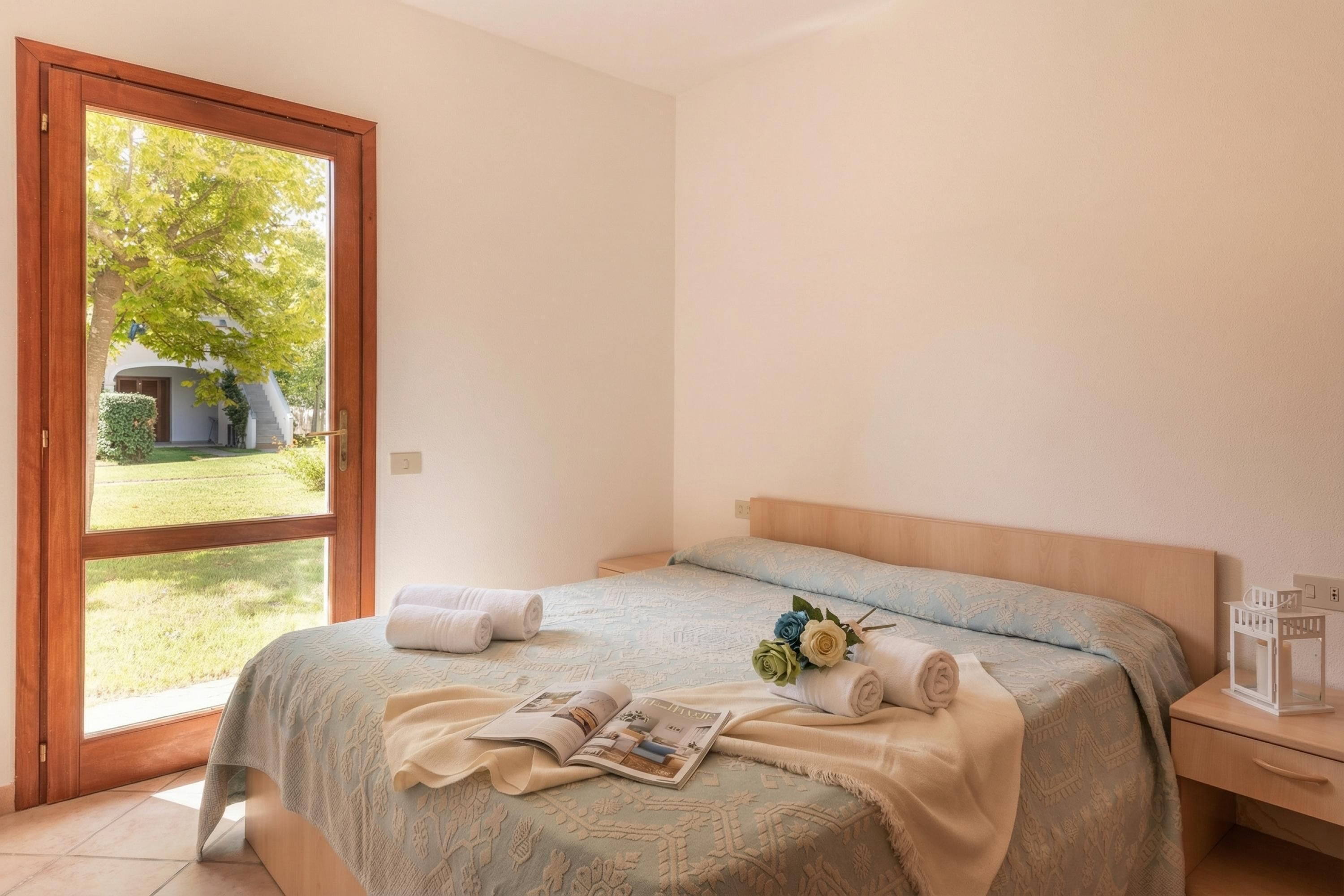 A bedroom with a bed, a nightstand, a window, and a view of the outdoors.
