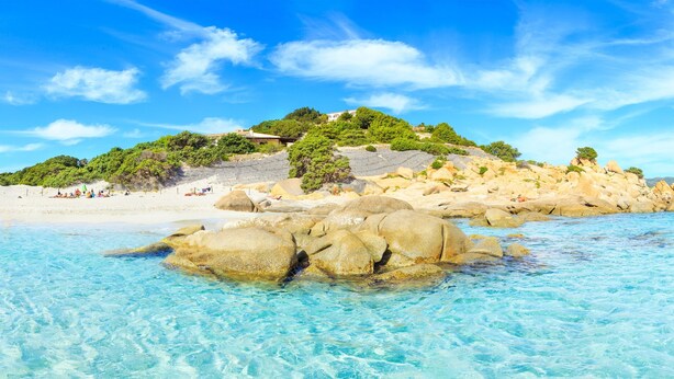 A beach with clear turquoise water, large rocks, and a hill with greenery and buildings in the background. - Le Fontane Bilo 4P 4 Km From The Beach, Villasimius, Italy (Villasimius)